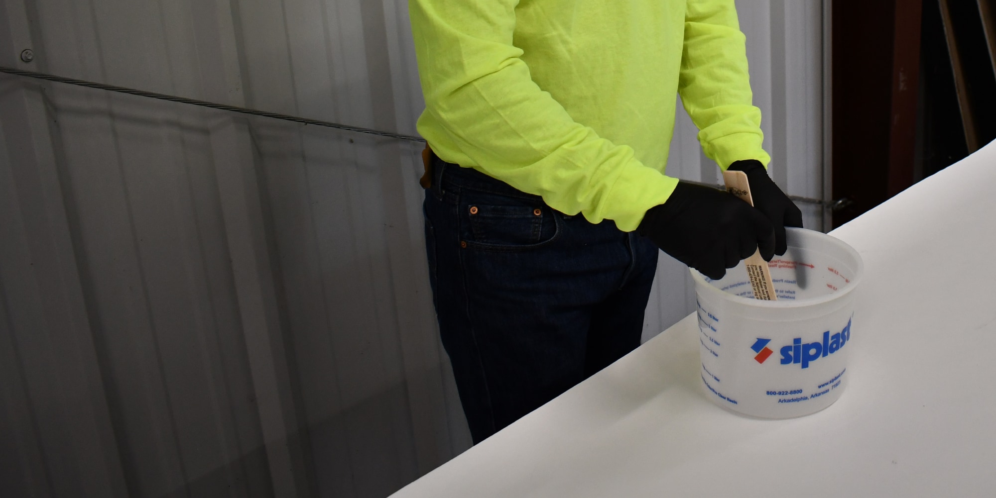 Siplast contractor wearing black gloves holding a Siplast bucket, mixing a Siplast product with a wooden tool